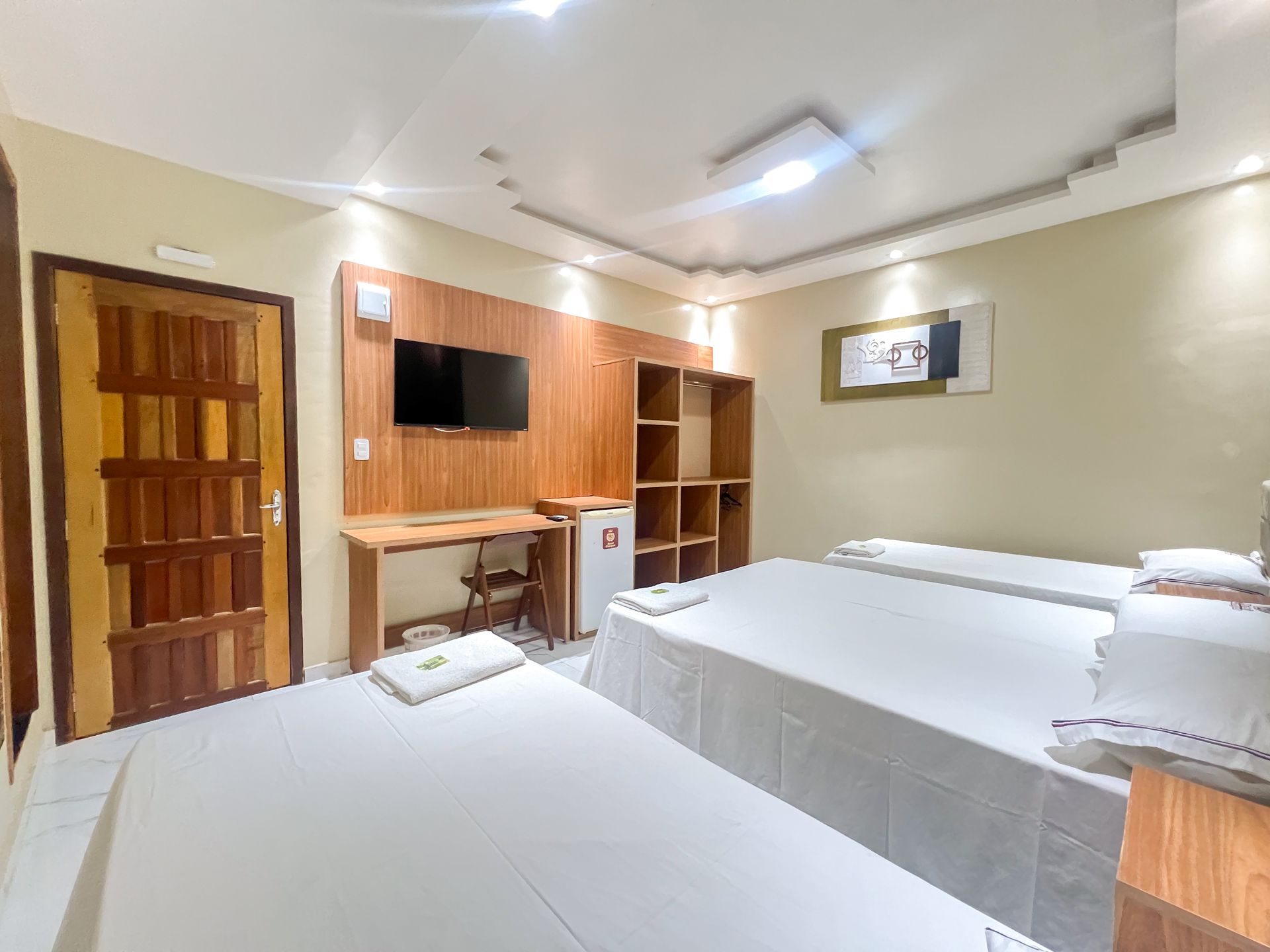 Quarto de hotel com duas camas, TV de parede, escrivaninha de madeira e paredes em tons creme aconchegantes.