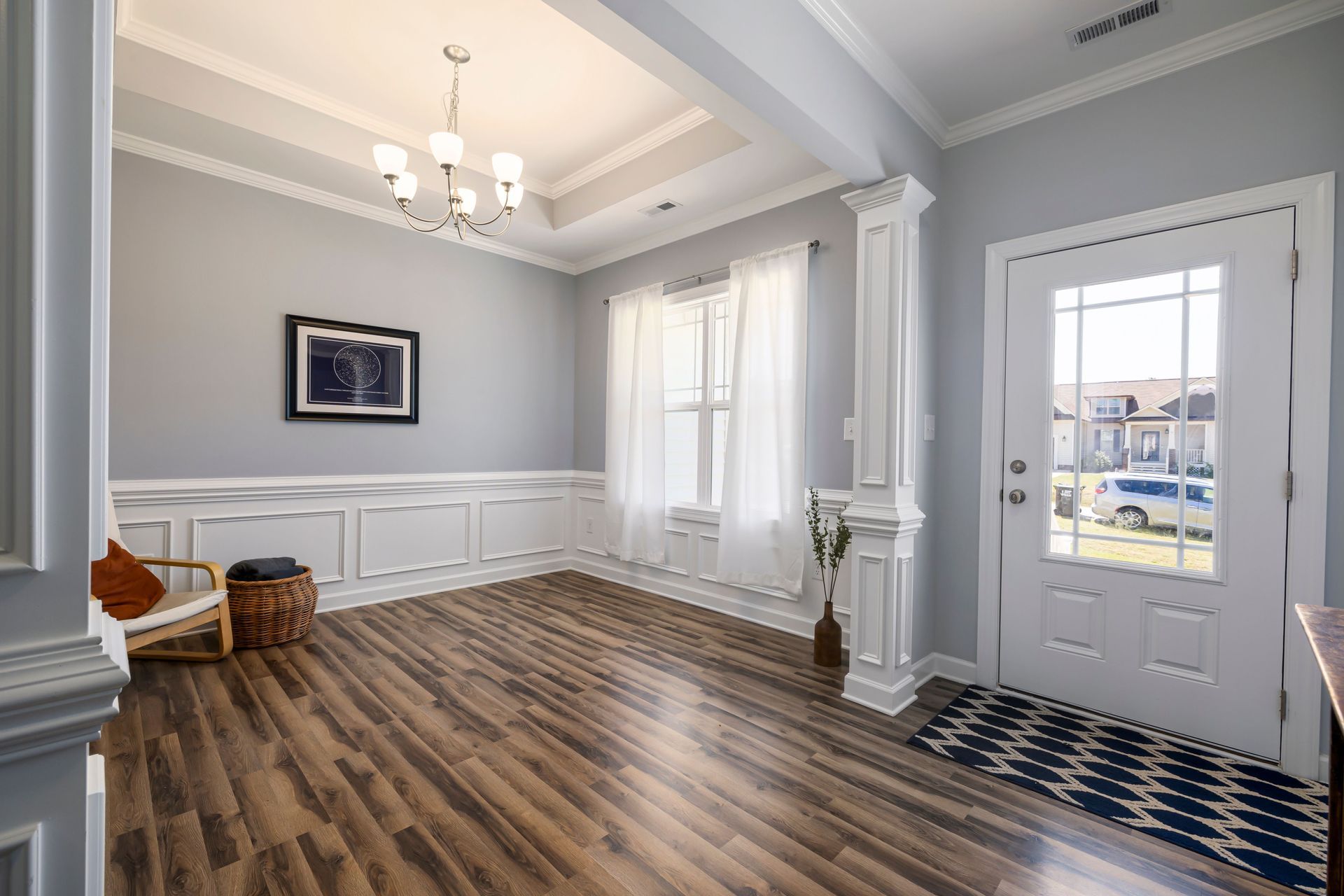 Empty living room with gray walls, wood floors, white trim, chandelier, and a glass-paneled door