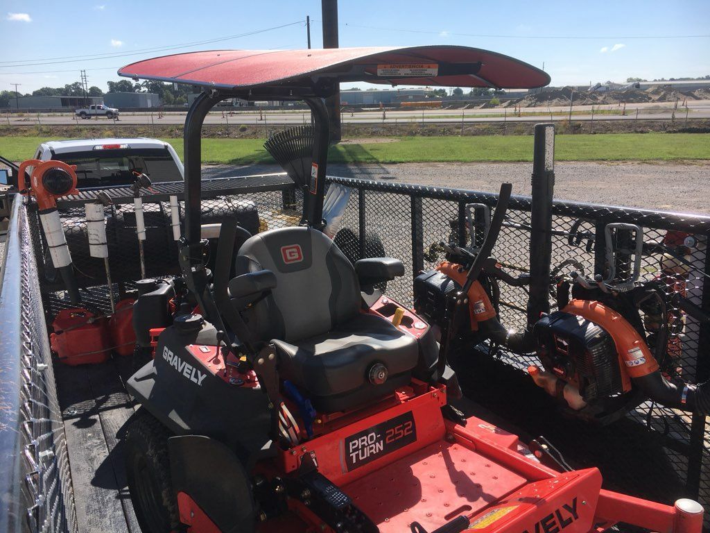 A red Gravely zero-turn mower with a shade canopy secured on a flatbed trailer, alongside landscaping tools.