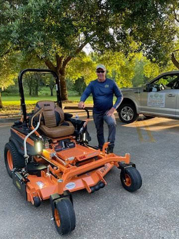 A person stands next to an orange zero-turn mower in a parking lot, with a truck parked in the background under trees.