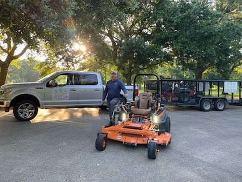 A person stands next to an orange commercial zero-turn mower in a parking lot, with a silver truck and trailer nearby.