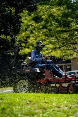 A person in a hat and blue work clothes drives a red and black Toro zero-turn mower across a grassy yard.