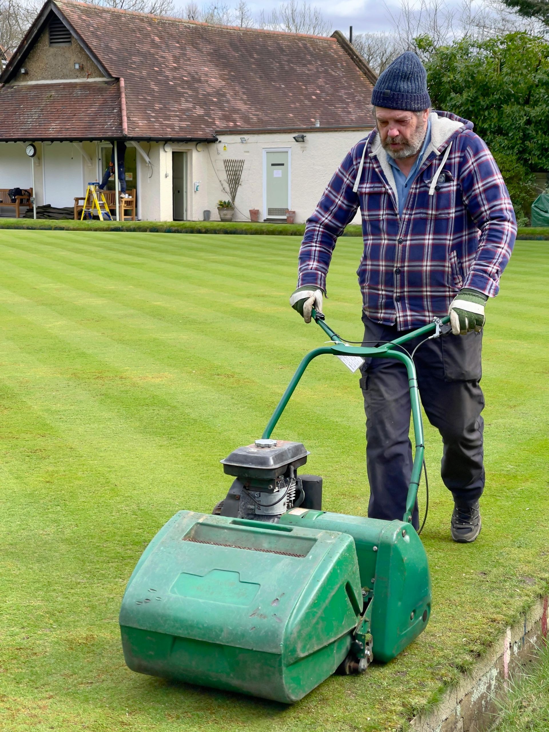 A person in a plaid jacket and beanie mows a manicured lawn with a green walk-behind mower in front of a building.