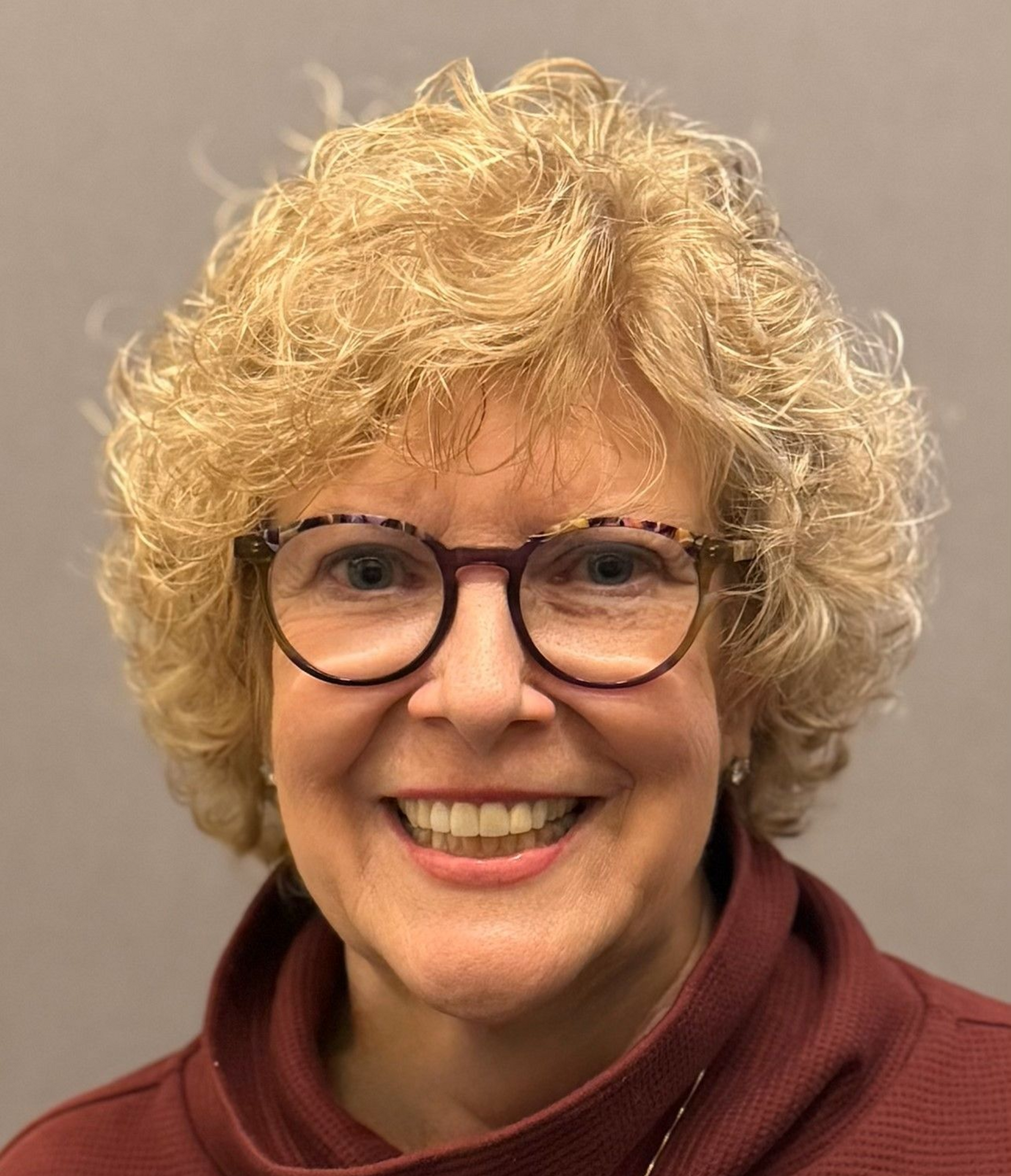 Smiling person with curly blond hair and glasses, wearing a maroon sweater against a gray background
