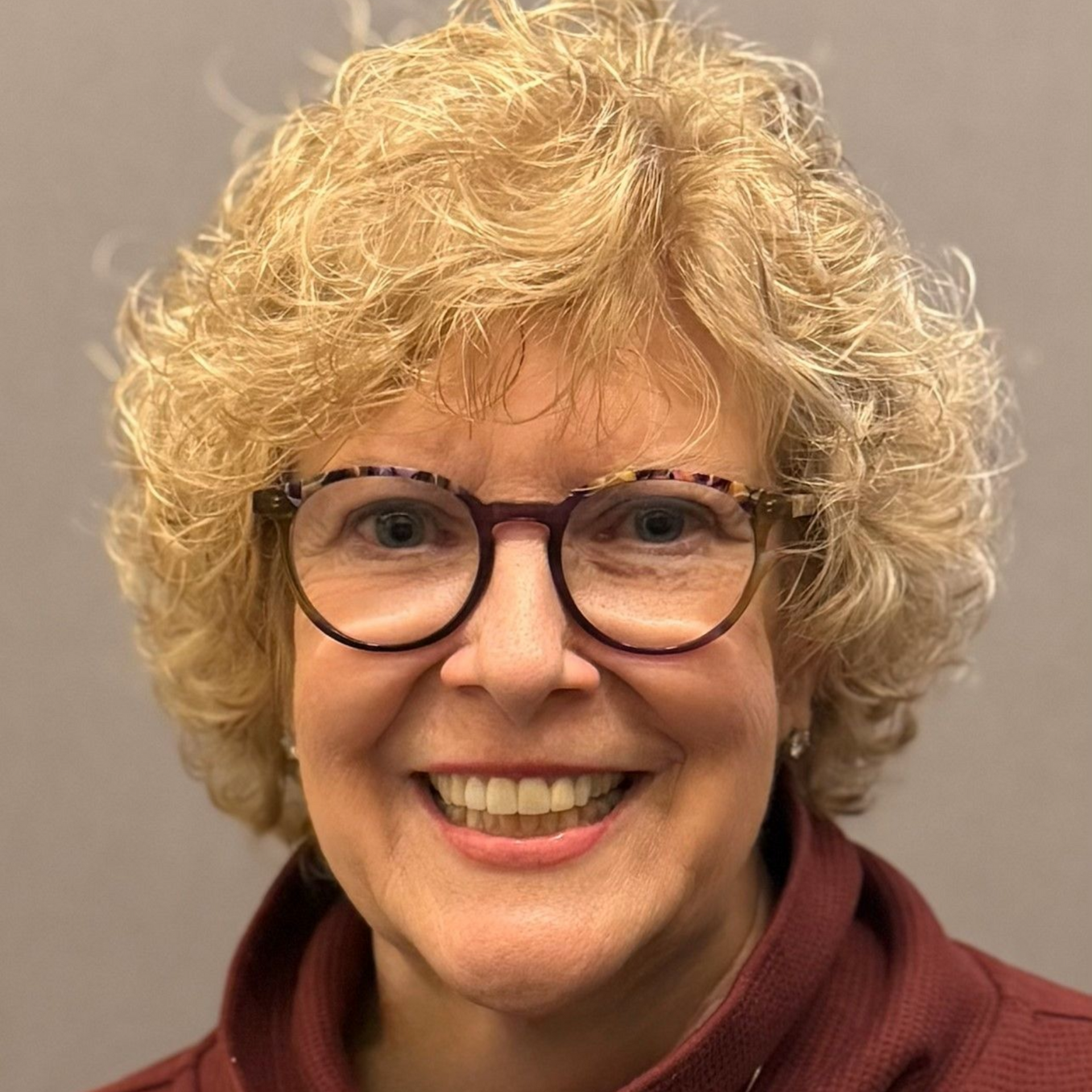Smiling person with curly blond hair and glasses, wearing a maroon sweater against a gray background