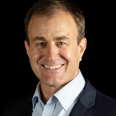 Smiling man in a light blue shirt and dark blazer against a black background