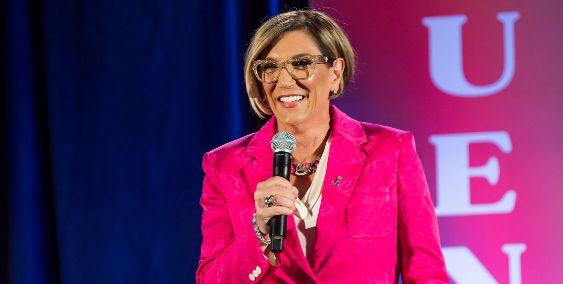 A woman in a pink jacket is holding a microphone and smiling.