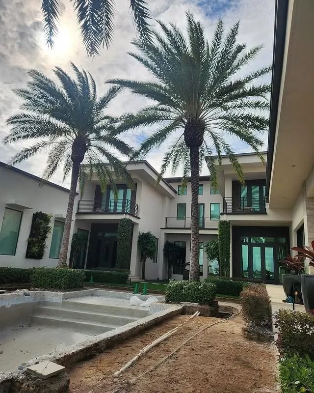 A modern multi-story home with large palm trees, green accents, and a pool under construction in the foreground.