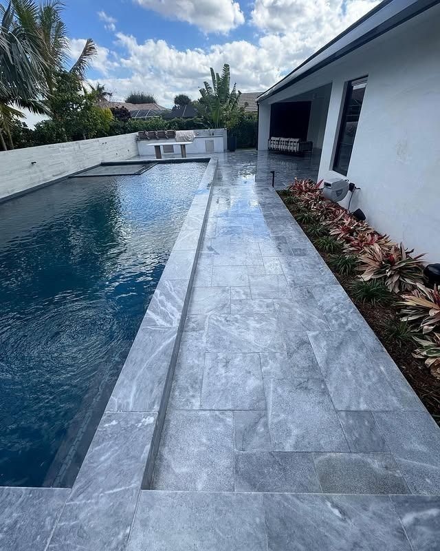 A swimming pool next to a white house with grey tiled patio flooring and garden beds under a bright, partly cloudy sky.