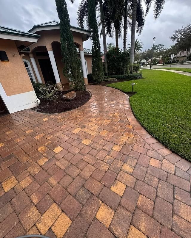 A curved paver walkway in shades of brown and tan leads to the front entrance of a stucco house with tall palm trees.