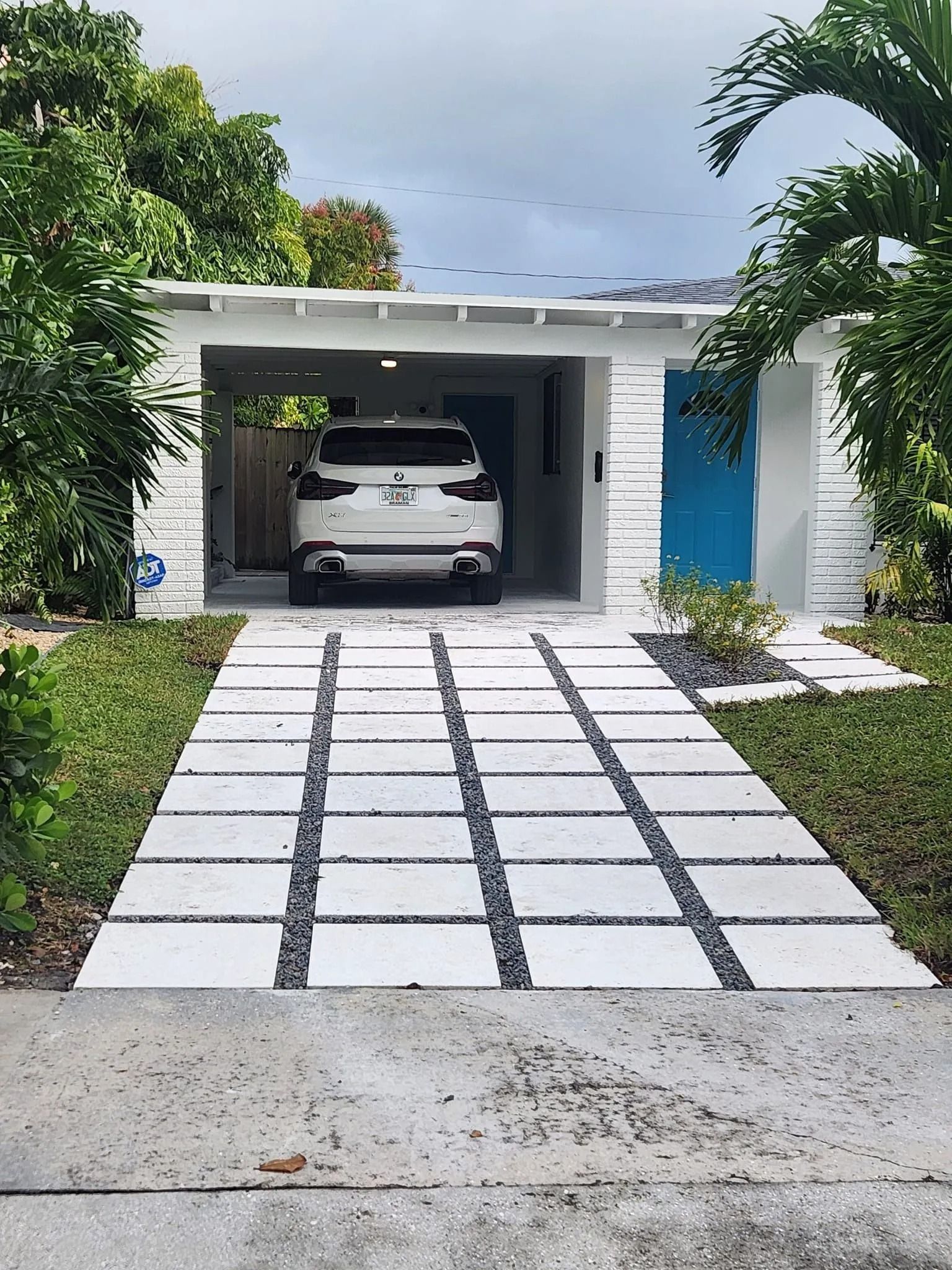 A white house with a blue front door and a carport, featuring a modern grid-patterned paver driveway leading to it.