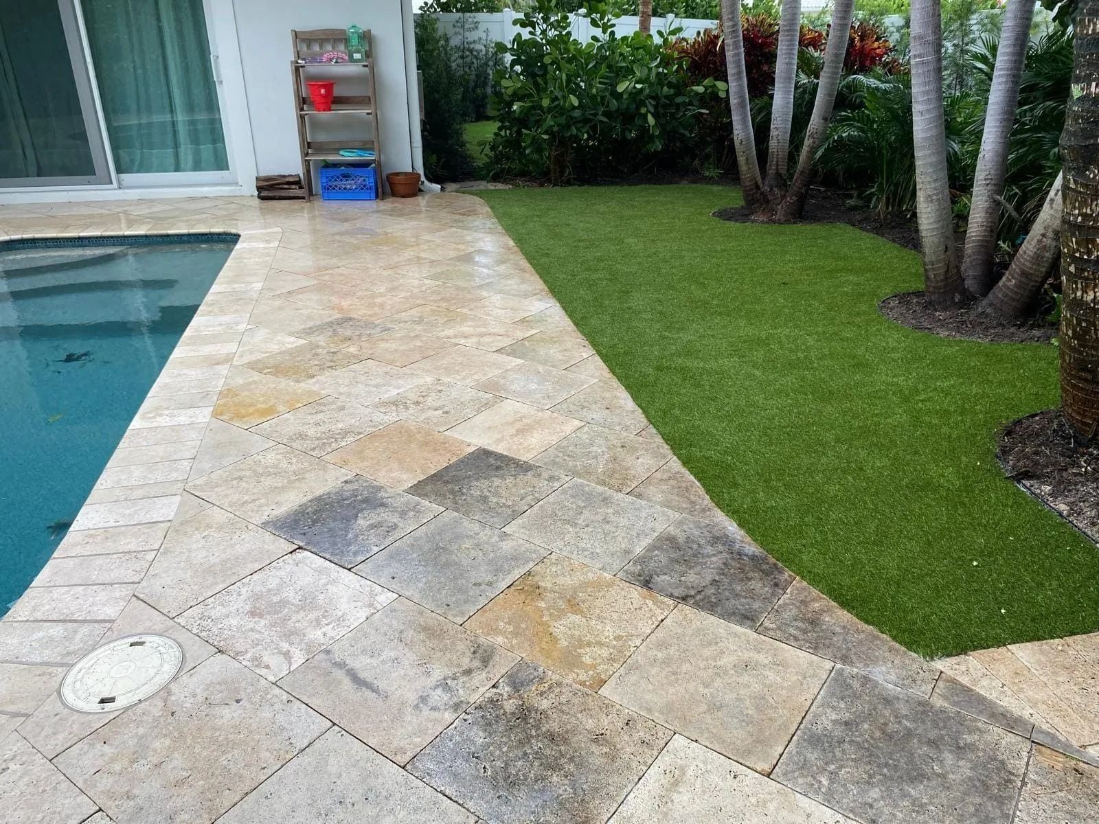 A backyard pool with beige stone patio tiles alongside a green lawn, tropical trees, and a house exterior.