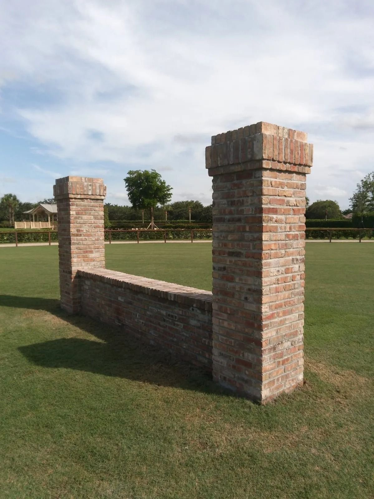 A low brick wall connecting two brick pillars stands in the middle of a grassy field under a bright, cloudy sky.