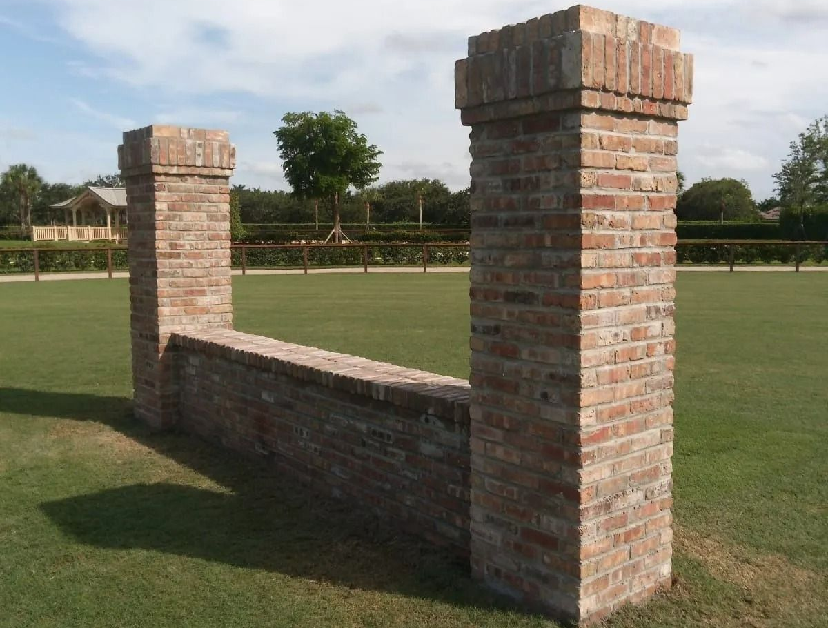 Two brick pillars connected by a low brick wall standing in a grassy field under a partly cloudy sky.