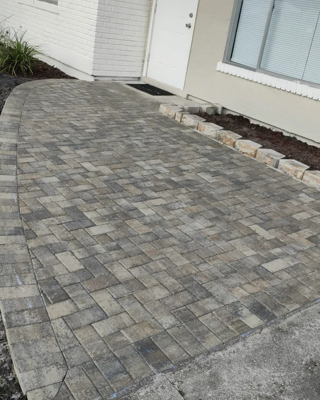 A curved paver walkway leading to a house entrance, bordered by stones and garden beds.