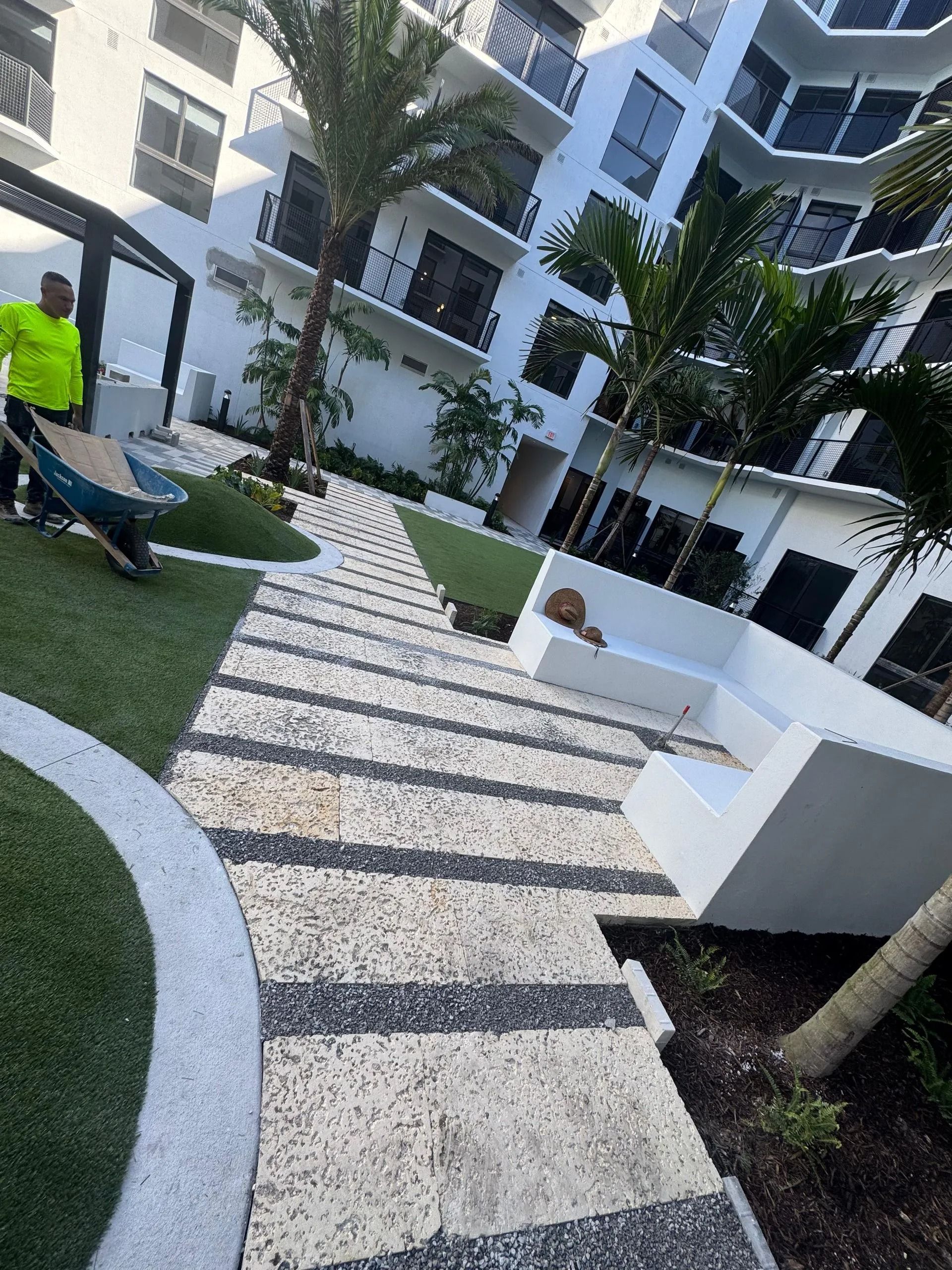 A worker with a wheelbarrow on a landscaped patio with stone pavers, artificial grass, palm trees, and a white bench.