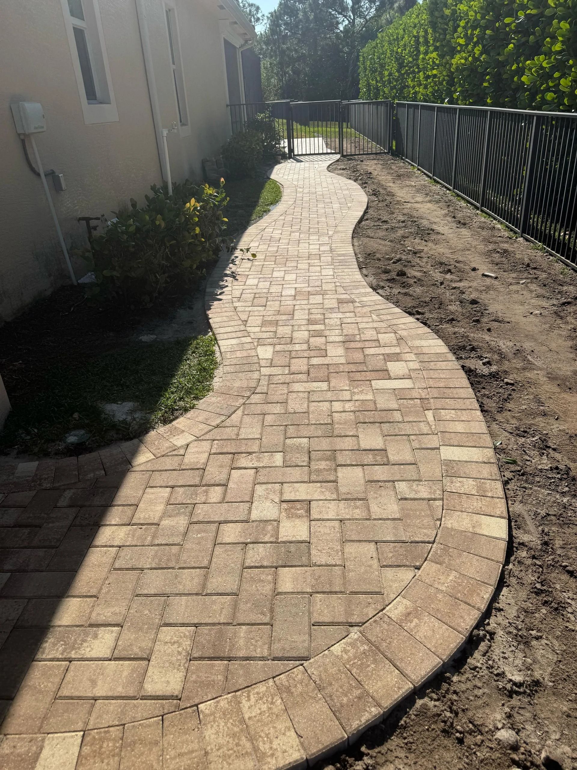 A curved brick paver walkway runs alongside a house and a dark decorative fence on a sunny day.