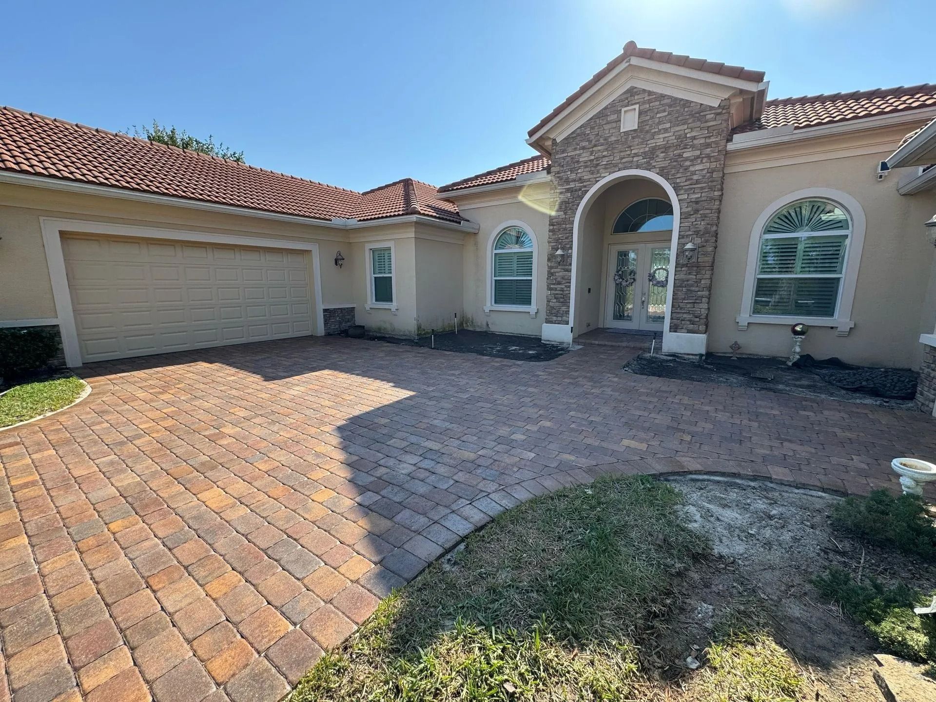 A single-story home with tan stucco, a stone-accented entryway, a tiled roof, and a paved brick driveway.