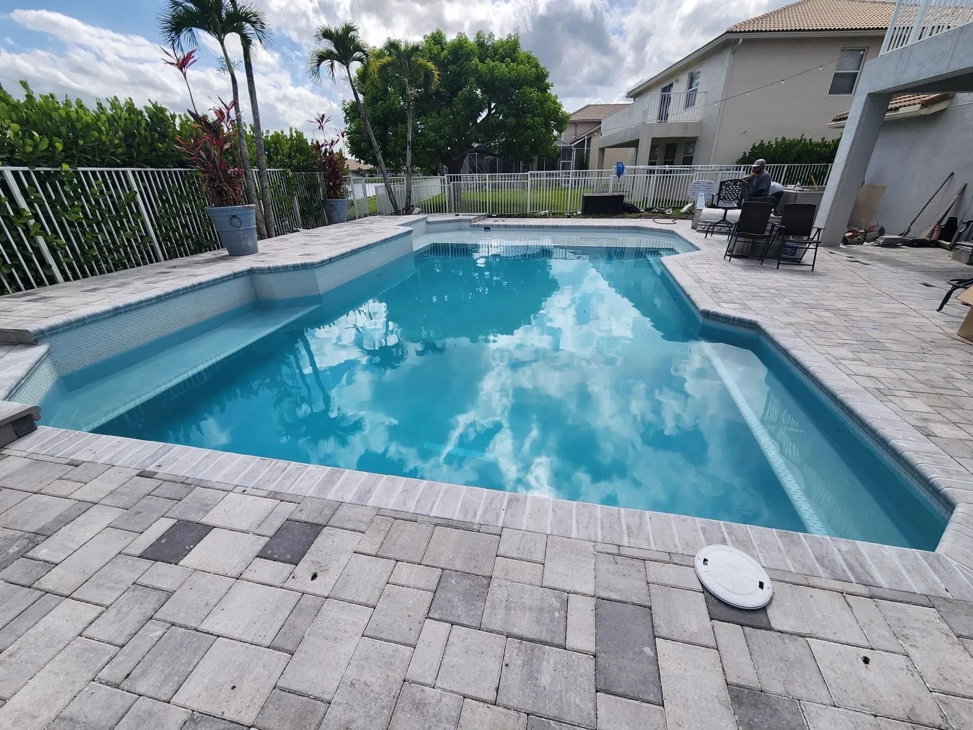 A backyard swimming pool with light blue water and stone paver decking, framed by a white fence and lush green trees.