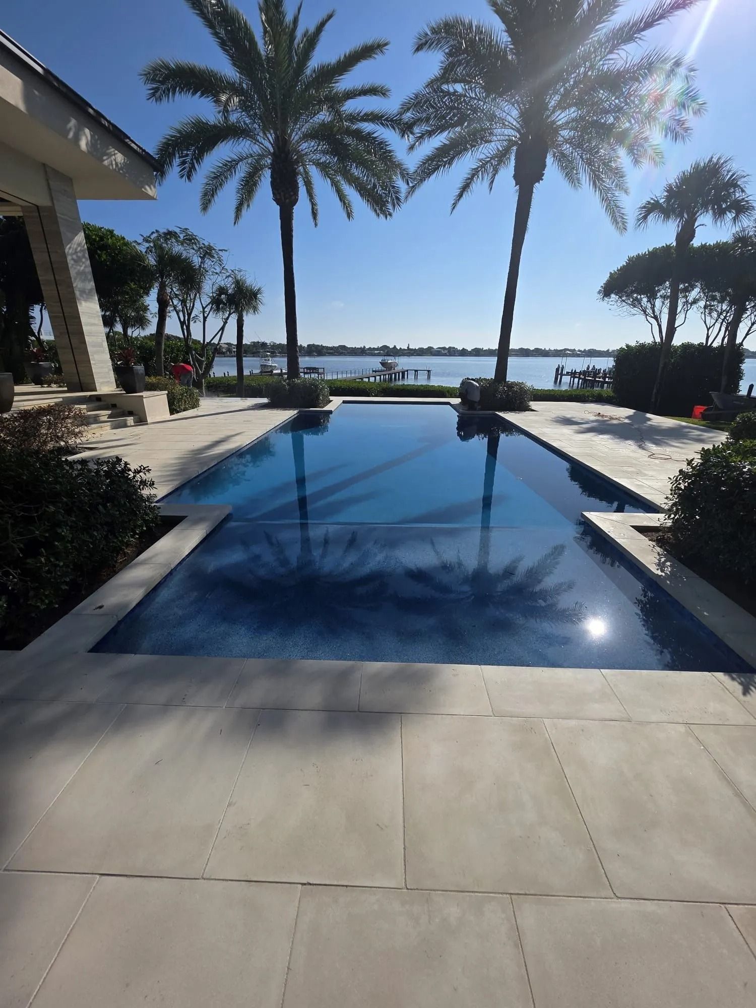 A rectangular swimming pool lined with palm trees overlooks a calm lake on a sunny day.