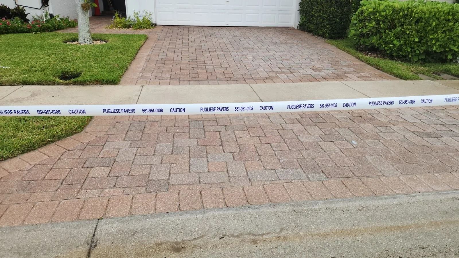 A white police barrier tape stretched across a brick driveway in front of a house, indicating a restricted area.