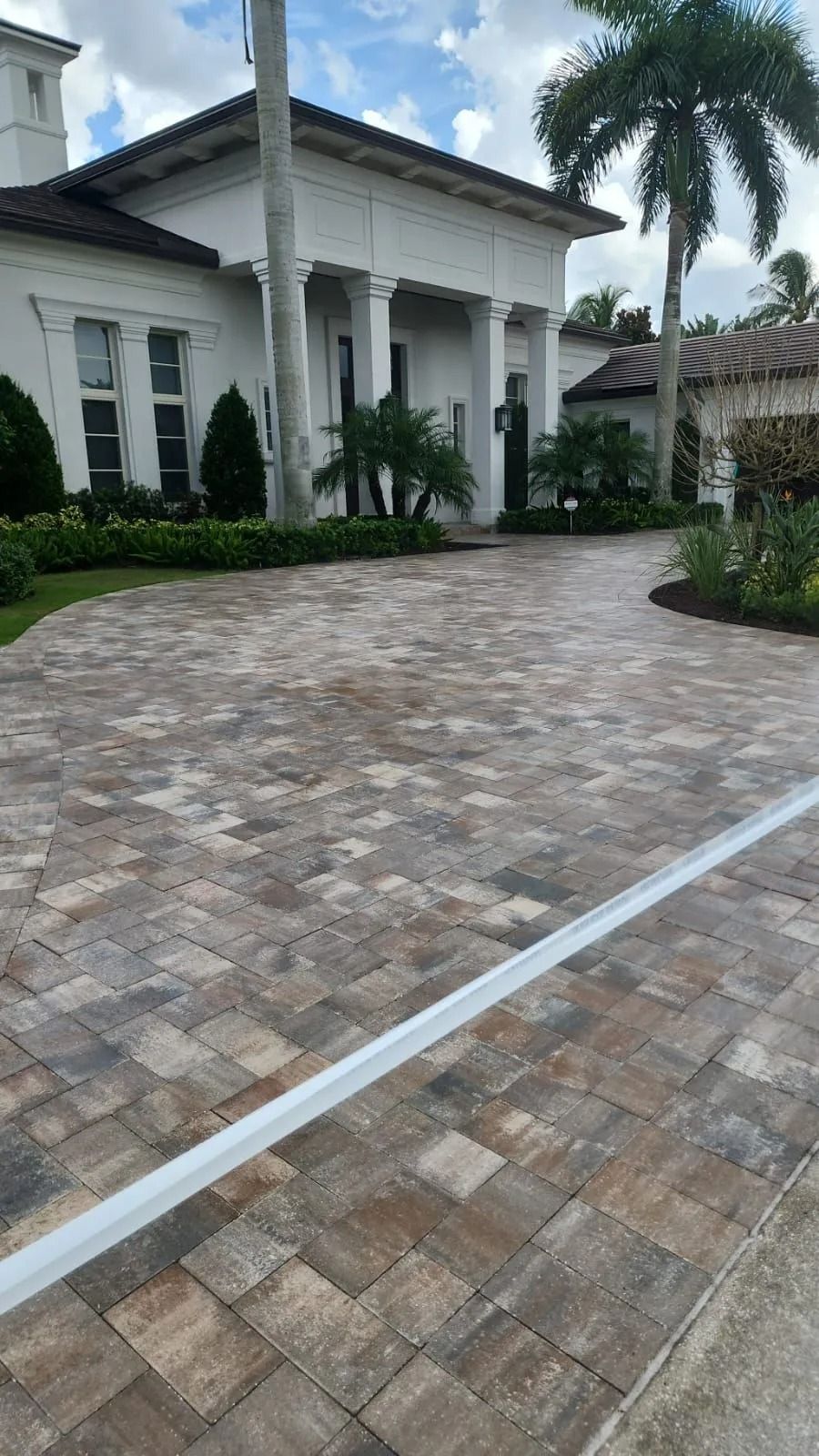 A white house with a multi-toned paver driveway, palm trees, and manicured green hedges under a blue sky.