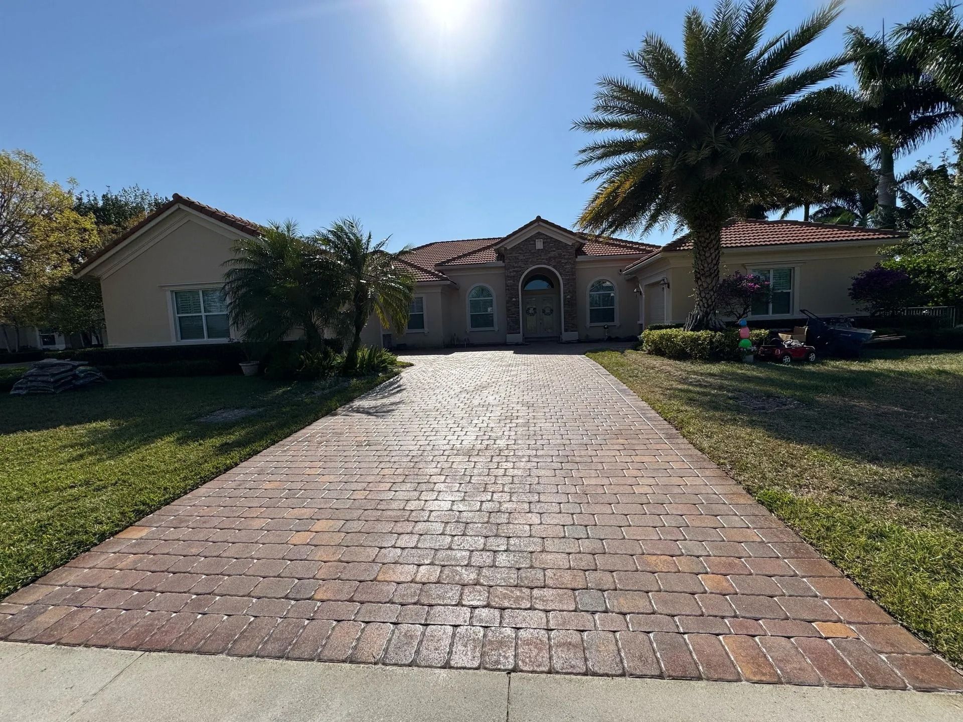 A wide, tan single-story house with a brick paver driveway, surrounded by green lawns and palm trees under a bright sun.