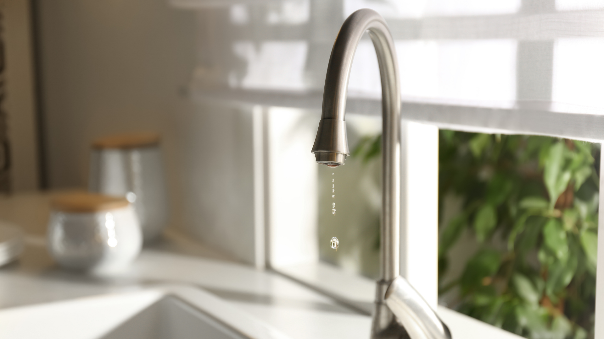 Faucet with dripping water in front of a window and blurry kitchen objects.