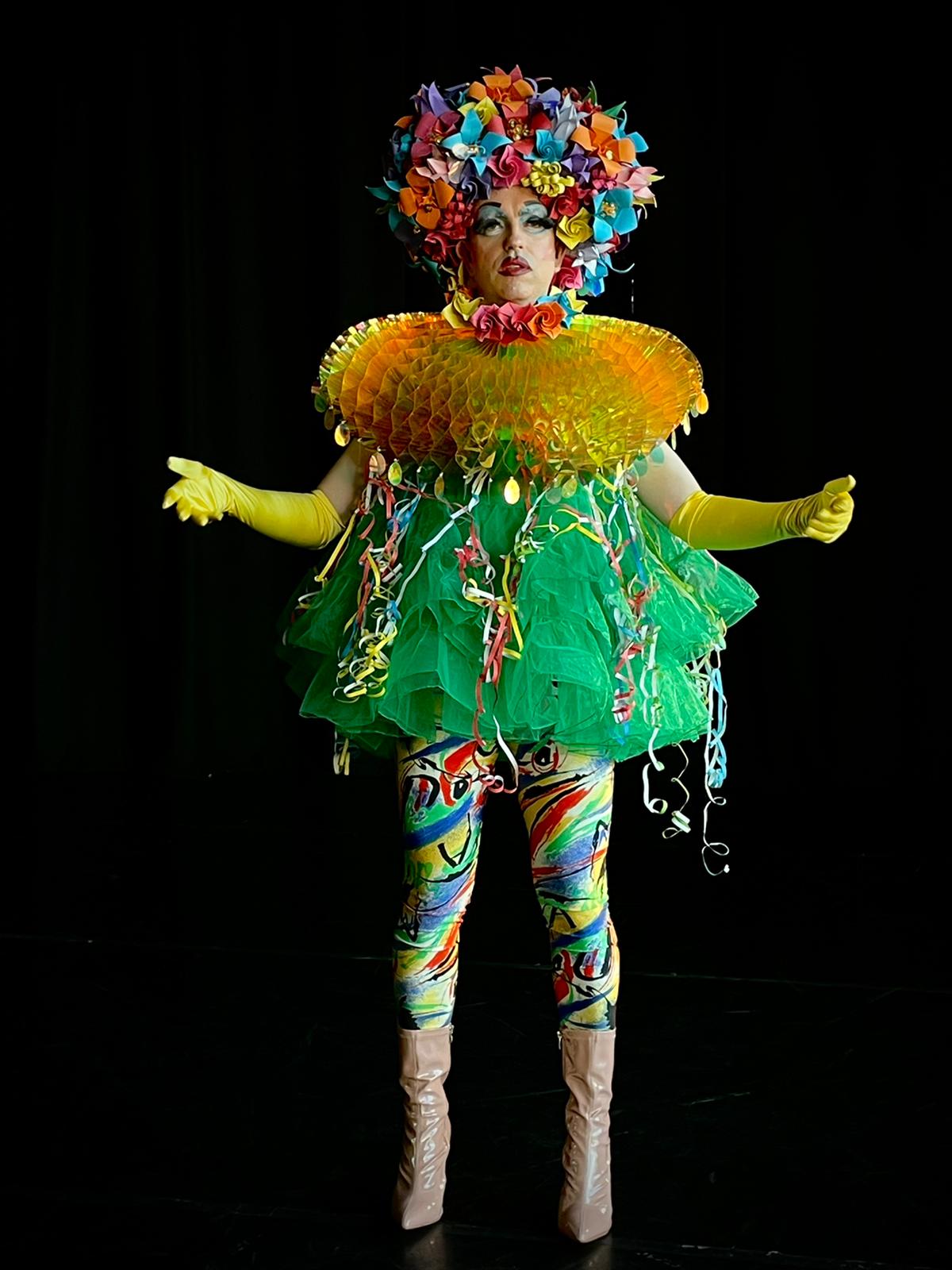 A man in a colourful costume and yellow gloves is standing on a black background.