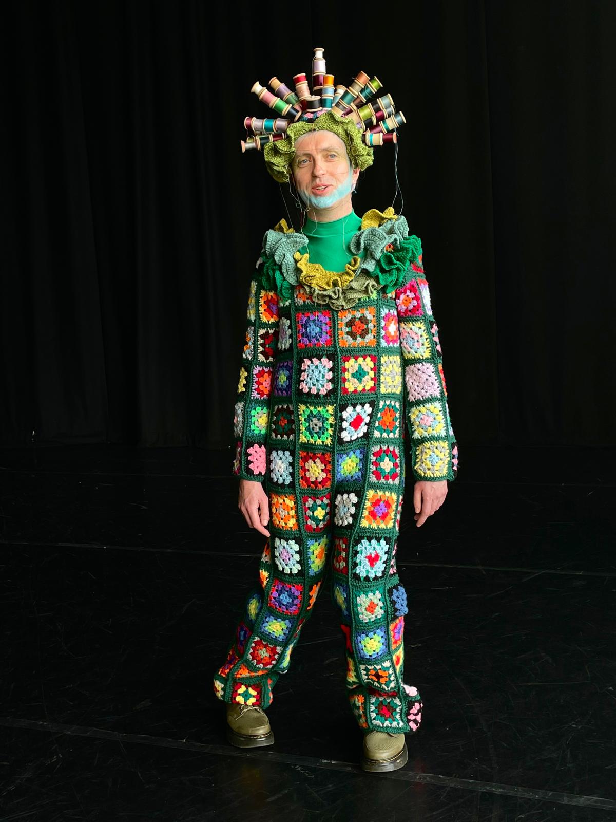 A man in a colorful crocheted outfit is standing on a stage.