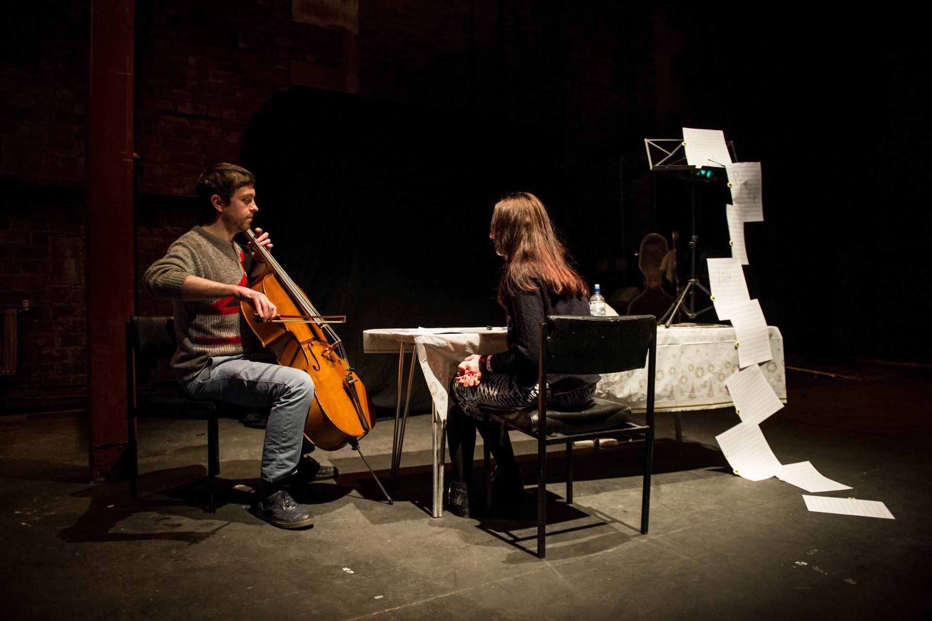 A man is playing a cello and a woman is sitting at a table.