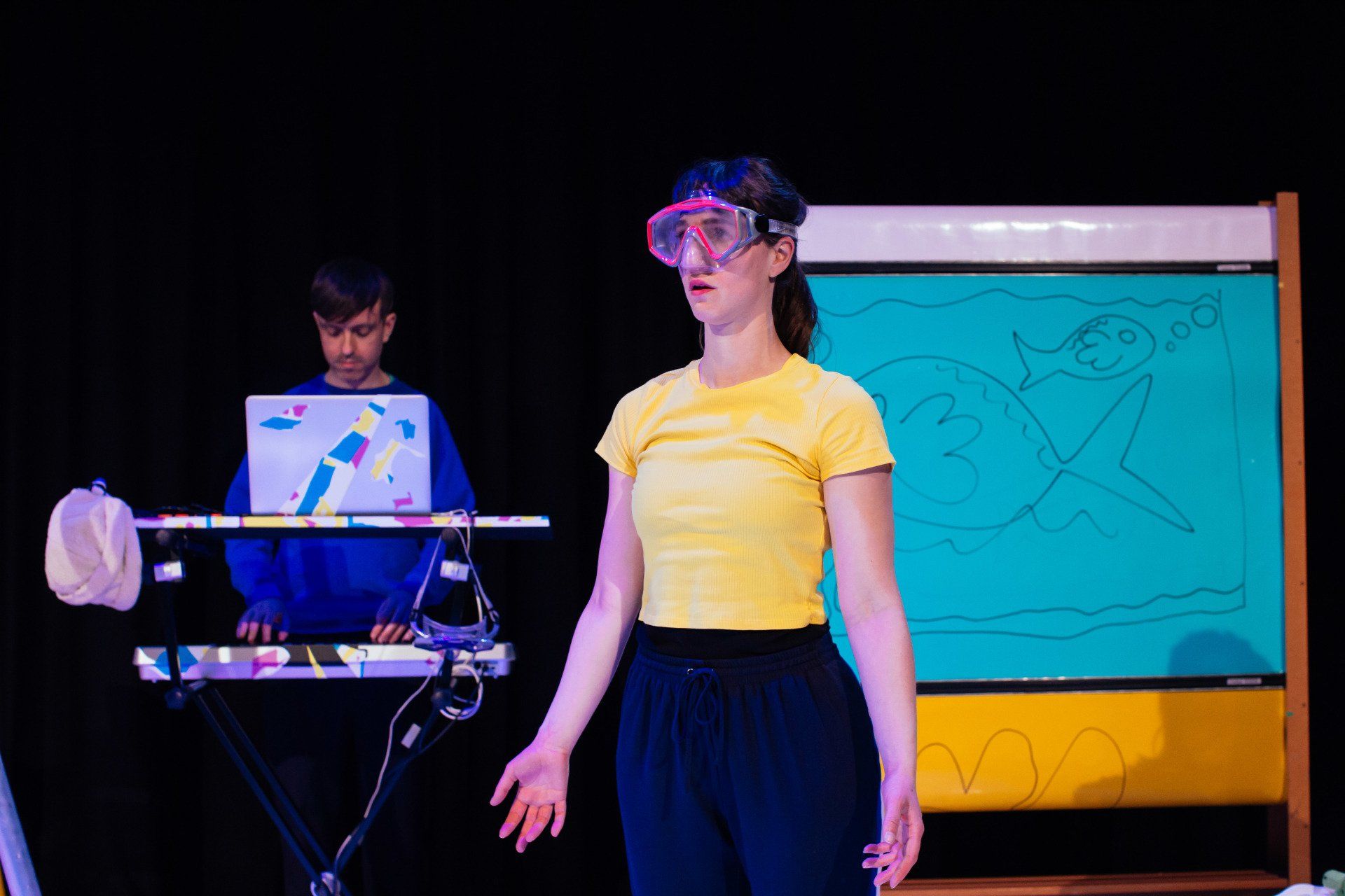 A woman wearing goggles is standing on a stage in front of a man playing a keyboard.