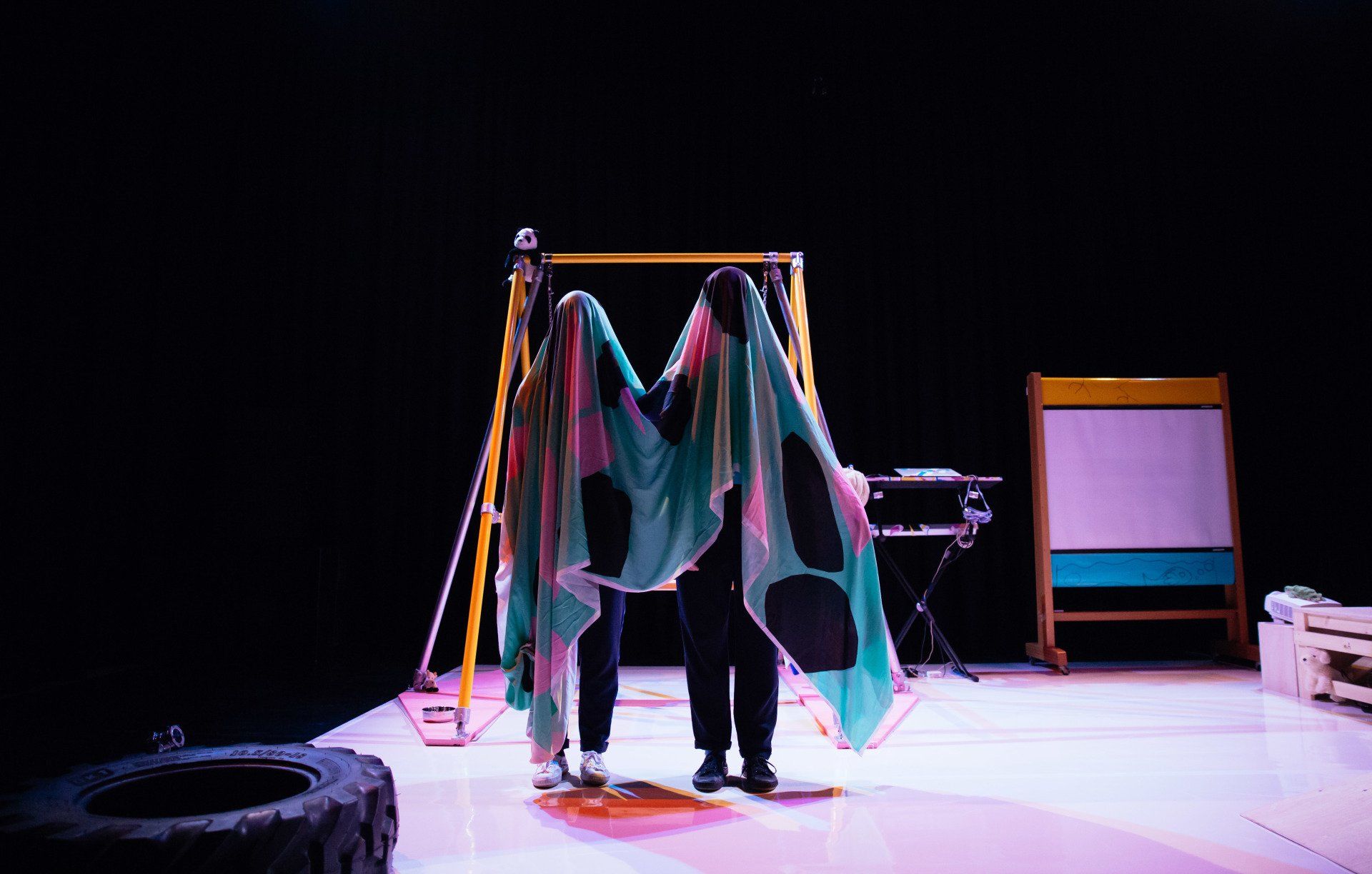 Two people are standing on a stage with a colourful cloth covering their faces.