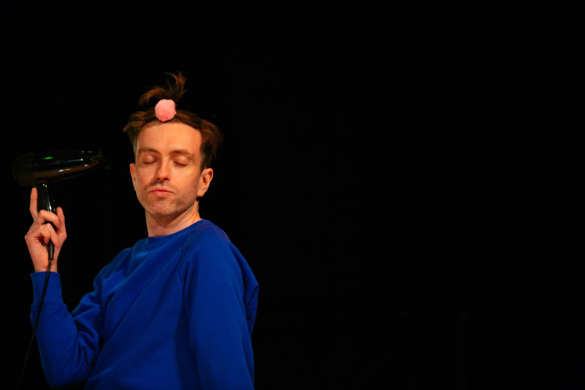 A man in a blue shirt is blow drying his hair with a pink ball on his head.