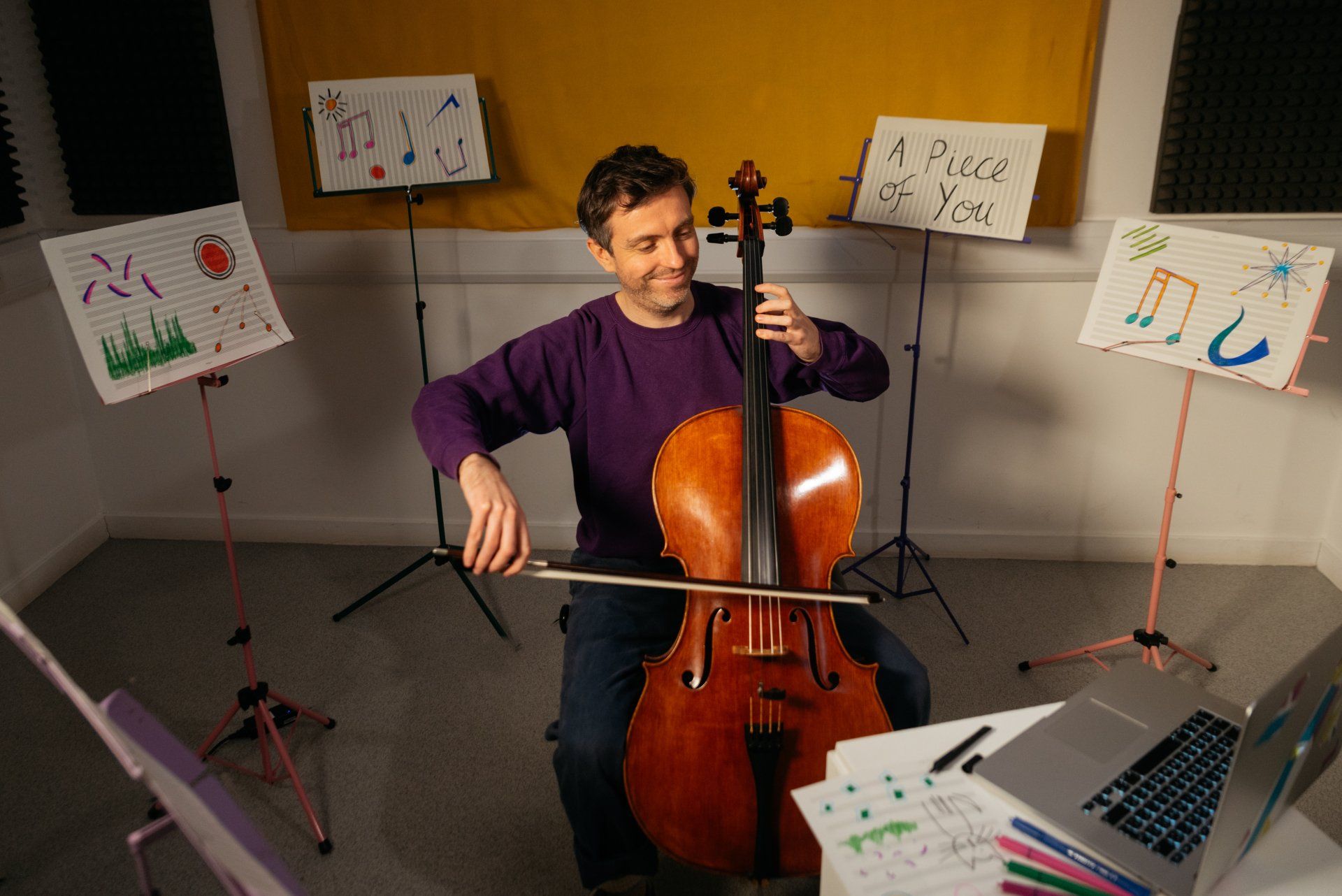 A man is playing a cello in a room with music stands and a laptop.