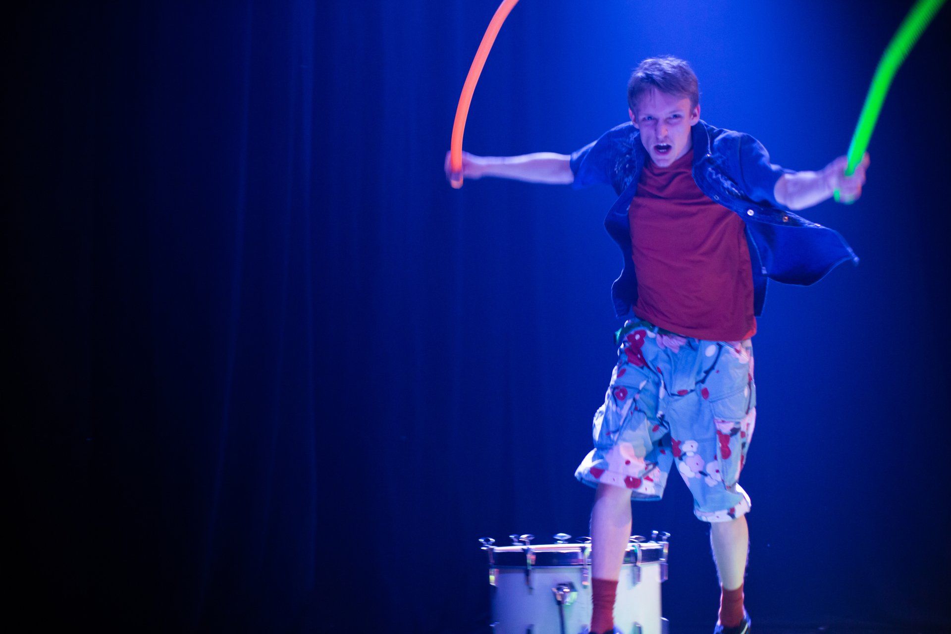 A man is playing drums on a stage while holding glow in the dark sticks.