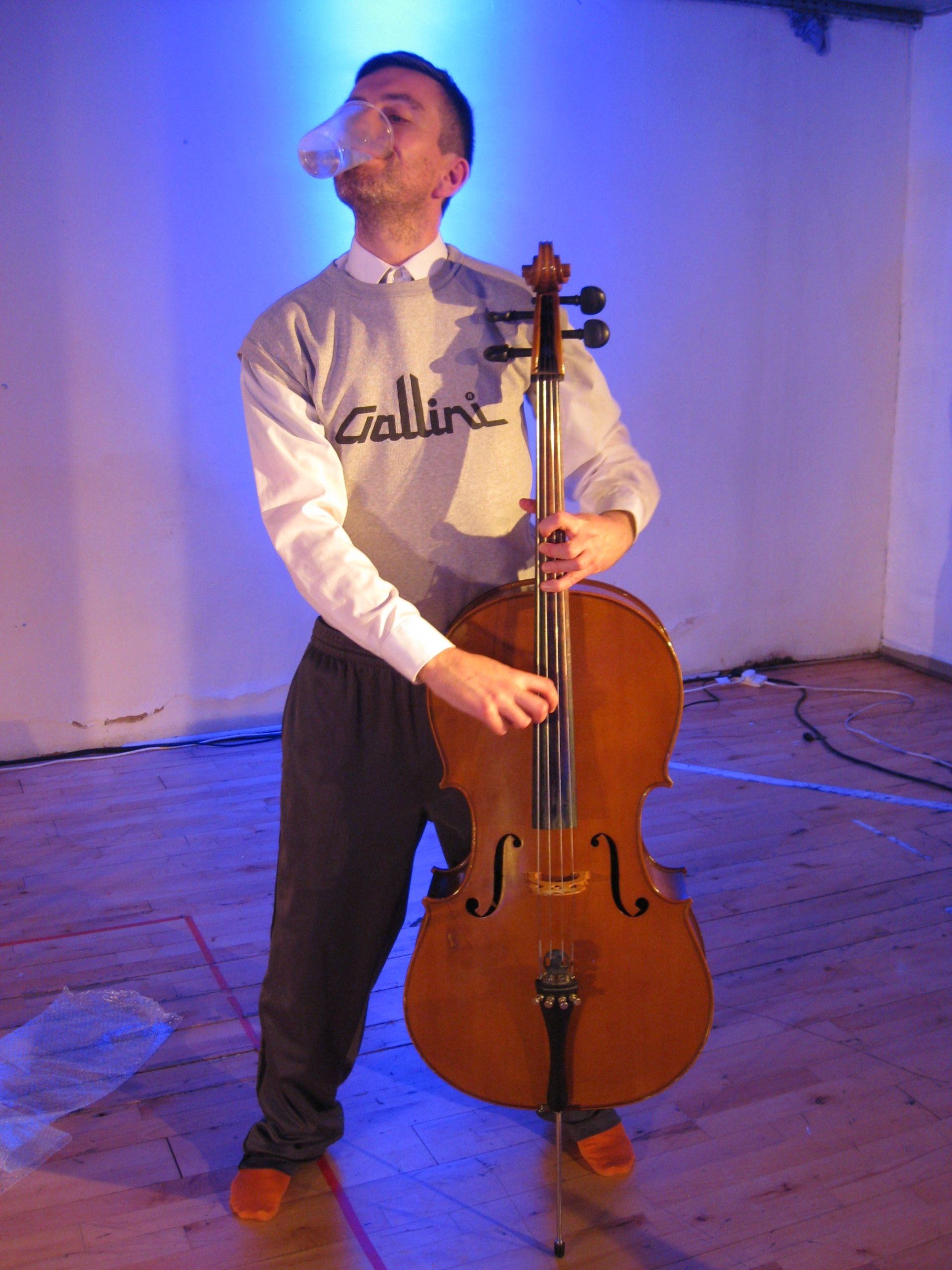 A man wearing a sweater that says collins is playing a cello with a glass of water held in his mouth
