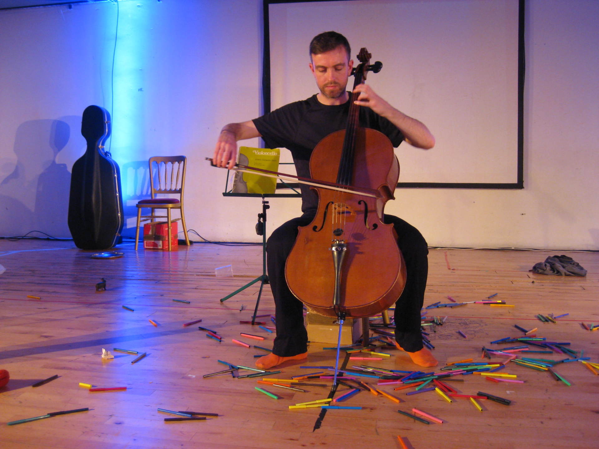 A man playing a cello on a stage surrounded by colouring pens.