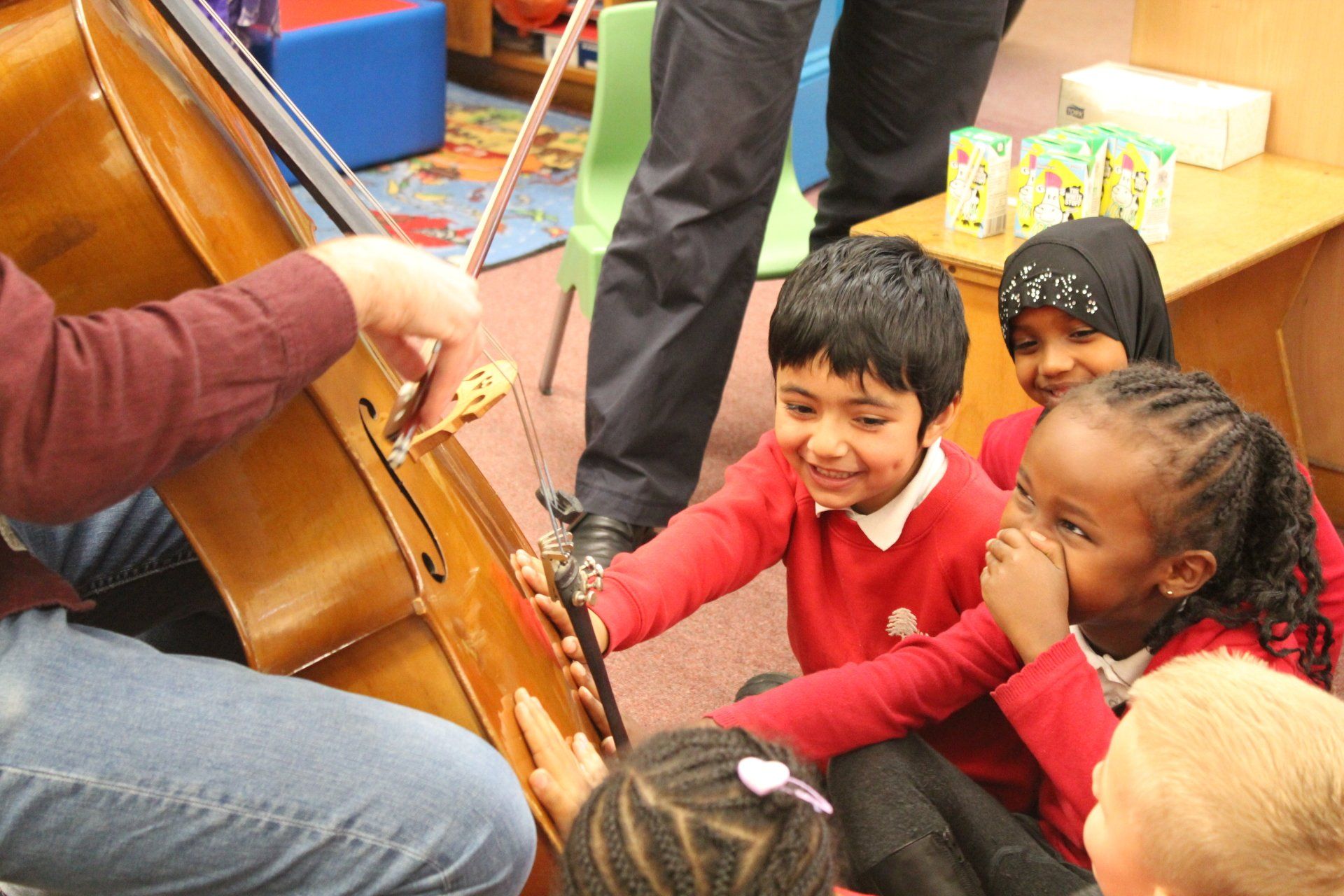 A group of children are sitting on the floor watching a man play a cello