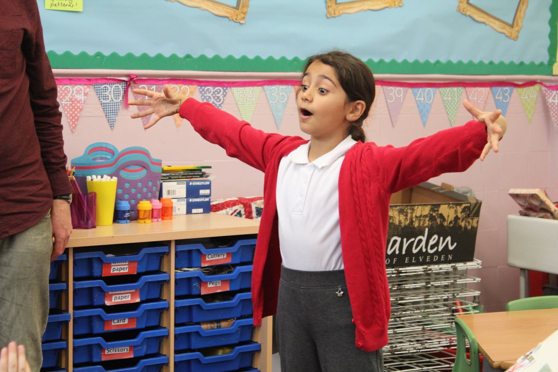 A girl in a red cardigan is standing in a classroom with her arms outstretched