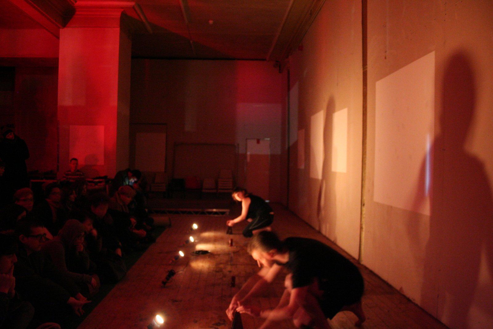 Two people are kneeling down in a dark room with torches and metronomes on the floor.