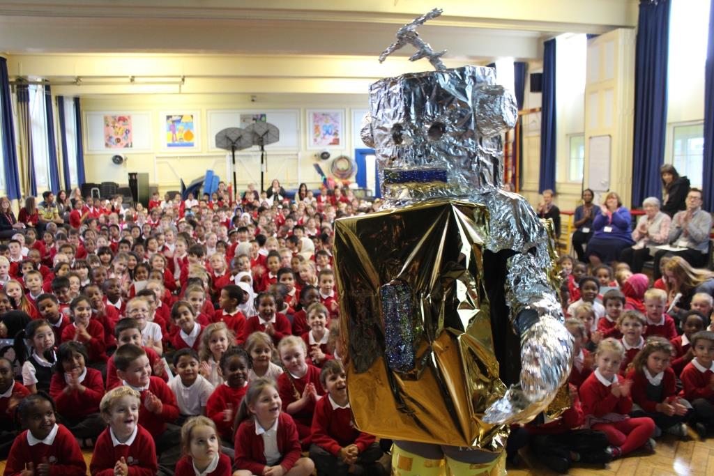Someone dressed as a robot stands in front of a large group of children sitting on the floor in a large room.