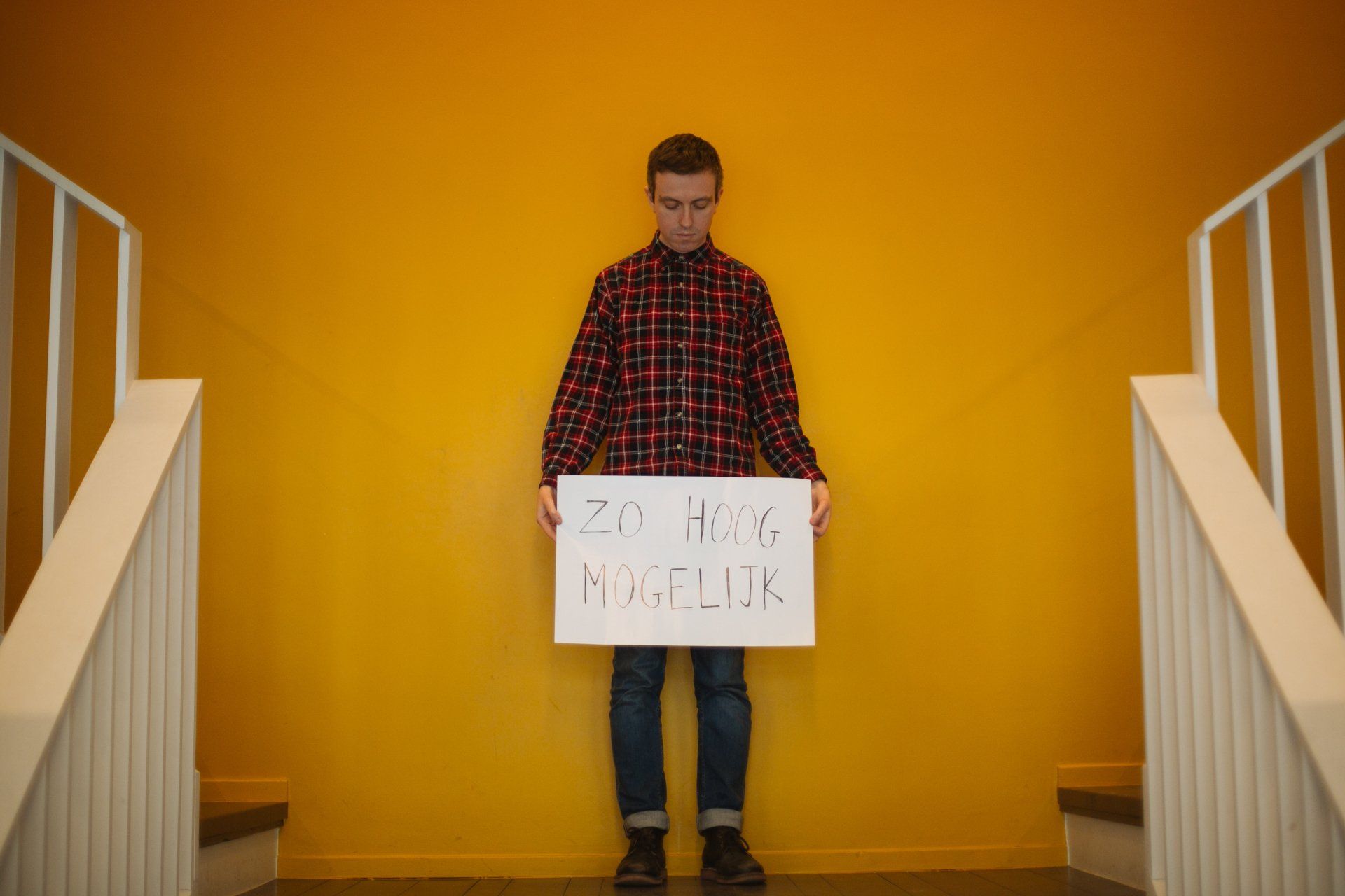 A man in a plaid shirt is holding a sign in front of a yellow wall.