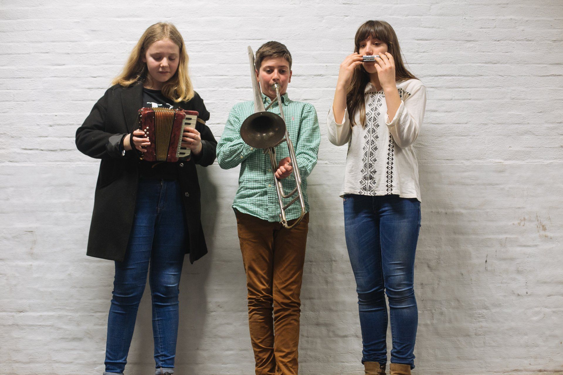 Three people are playing musical instruments in front of a white brick wall.