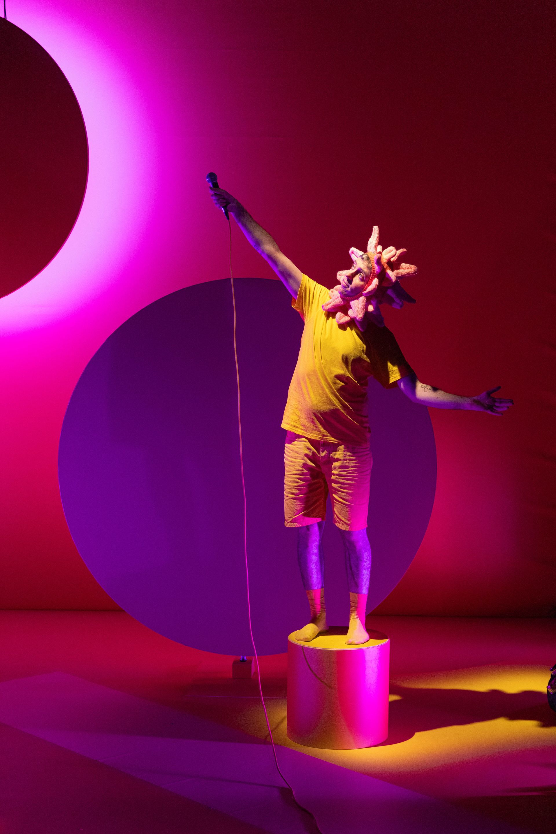 A performer wearing a pink headdress is standing with their arms outstretched.