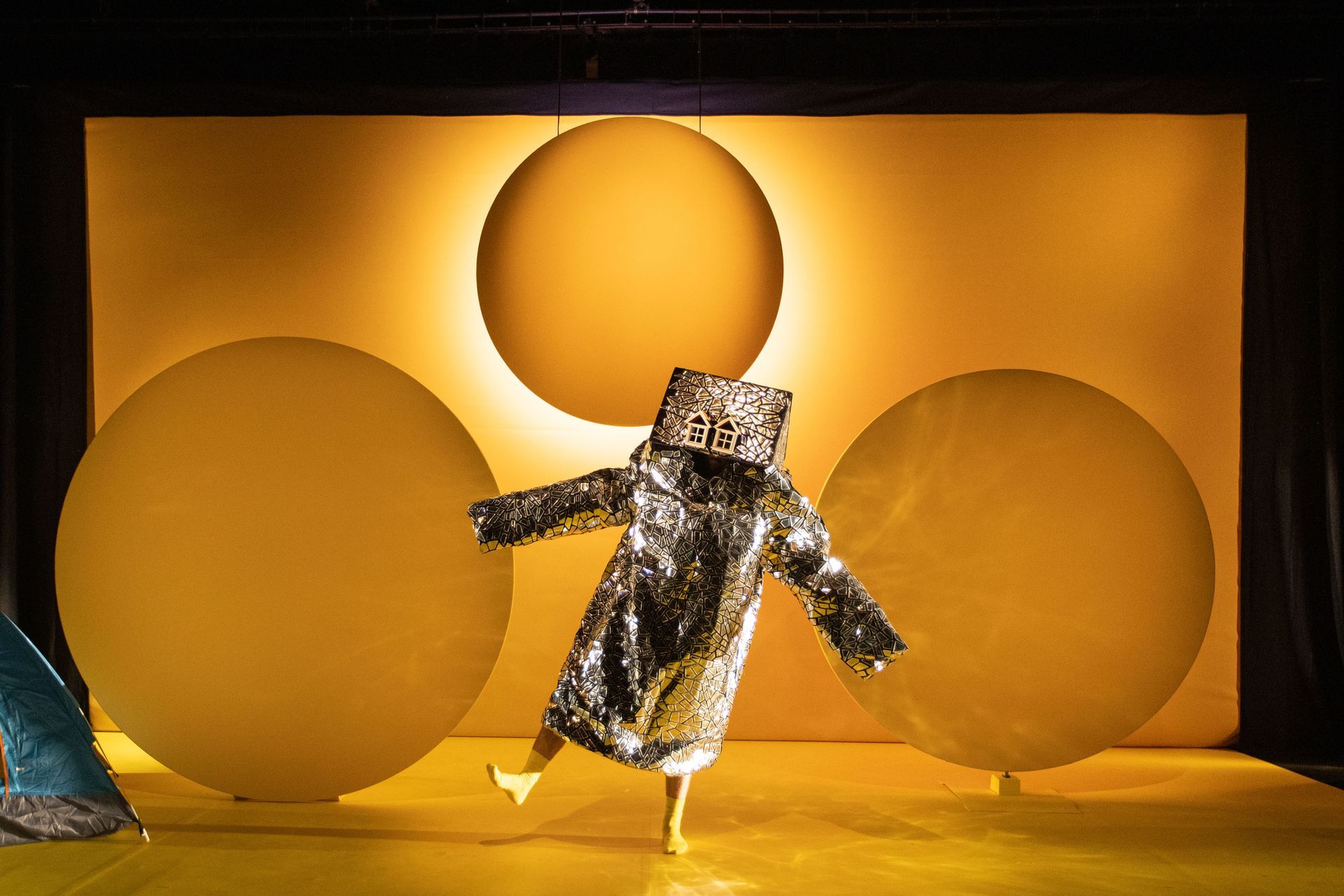A person in a silver doll’s house costume is dancing on a stage in front of a yellow background.