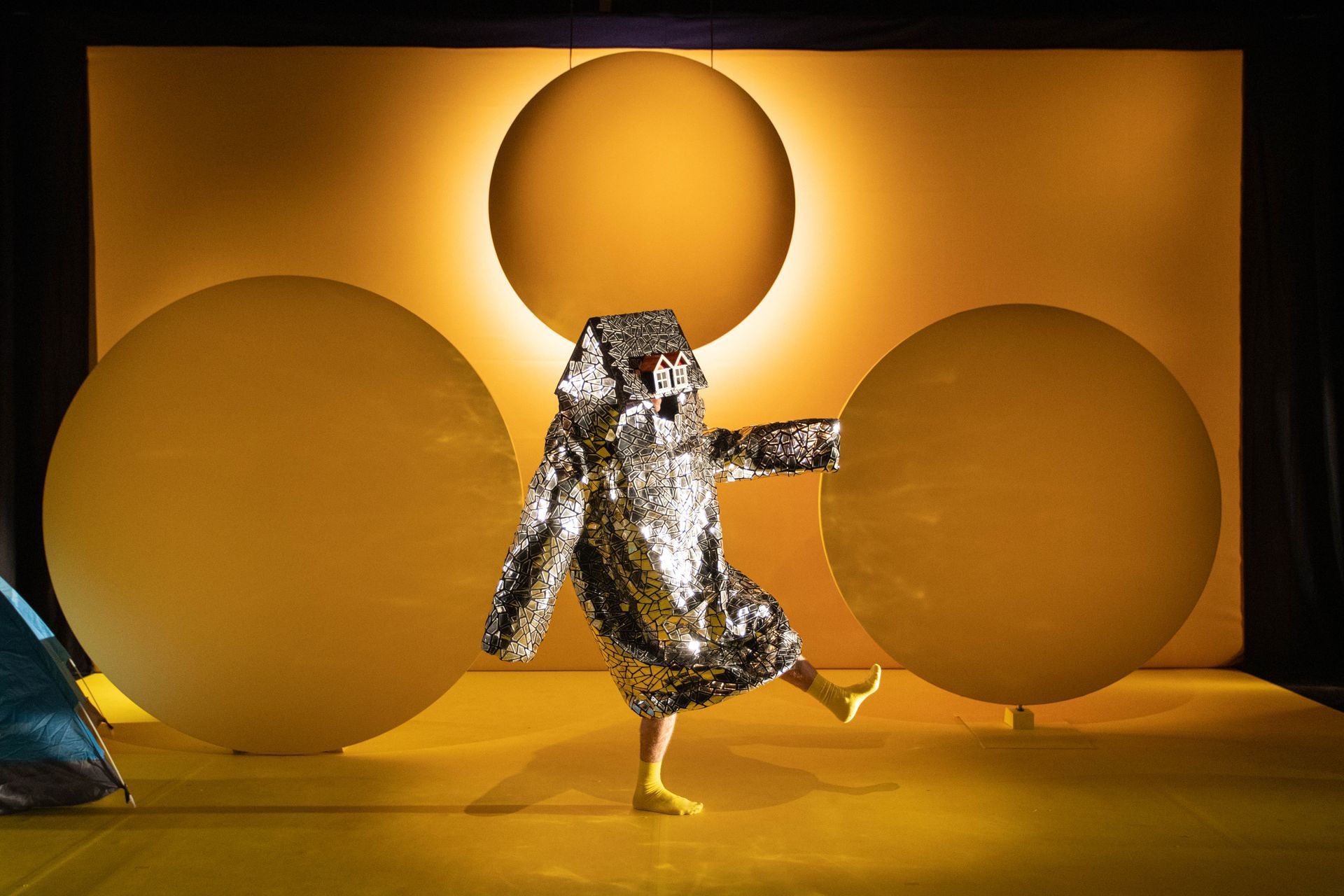 A person in a silver doll’s house costume is standing in front of a yellow background.