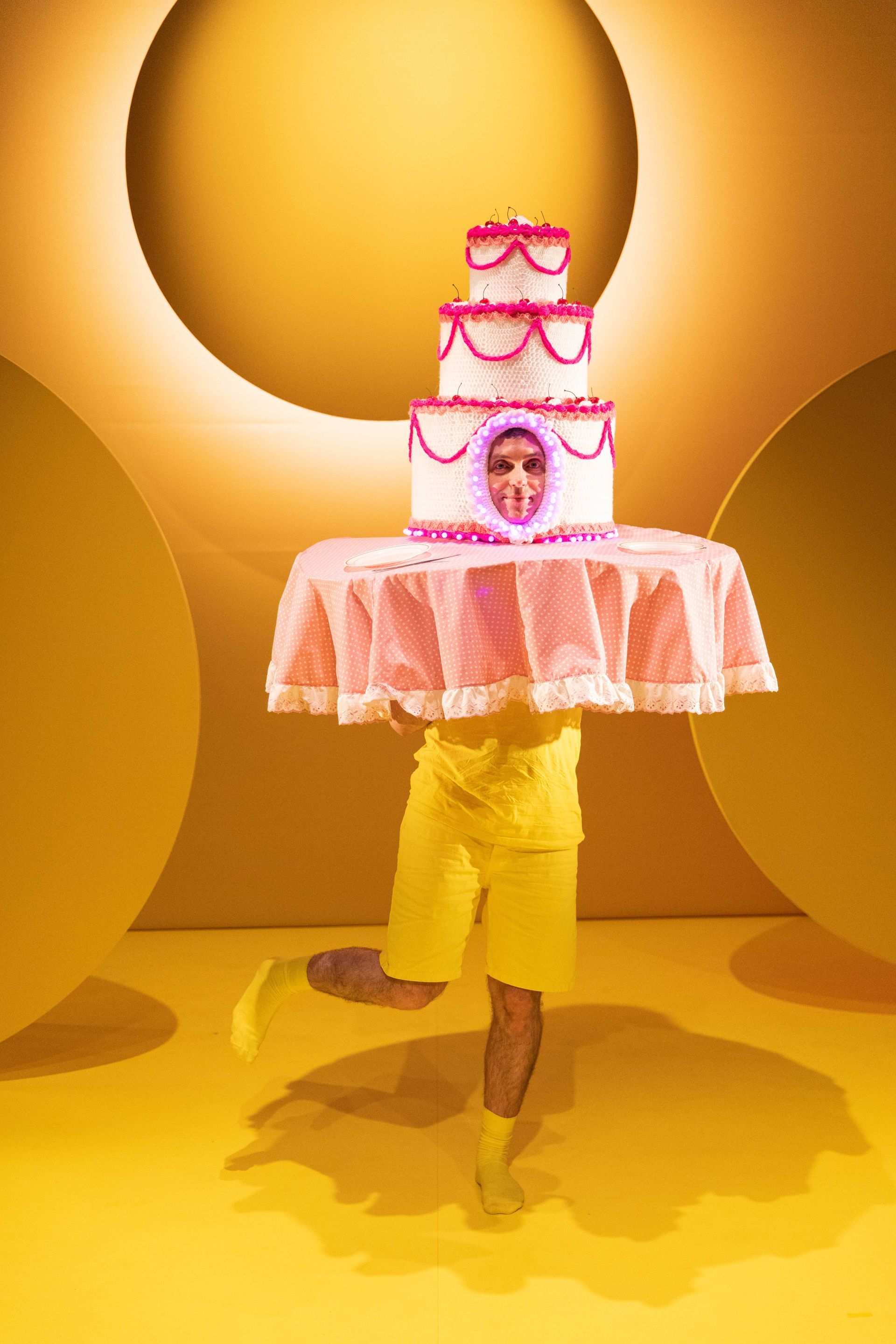 A man in yellow shorts is holding a cake costume on his head.