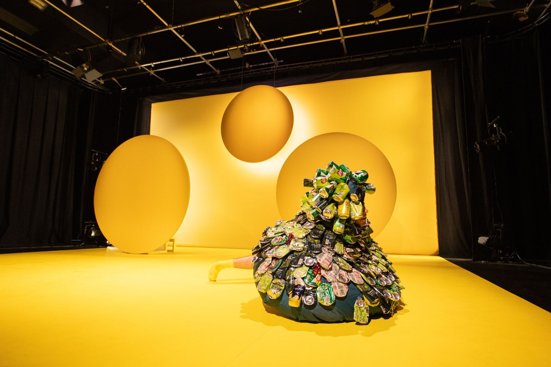 A sculptural bush-like costume is sitting on a yellow floor in front of a yellow wall.
