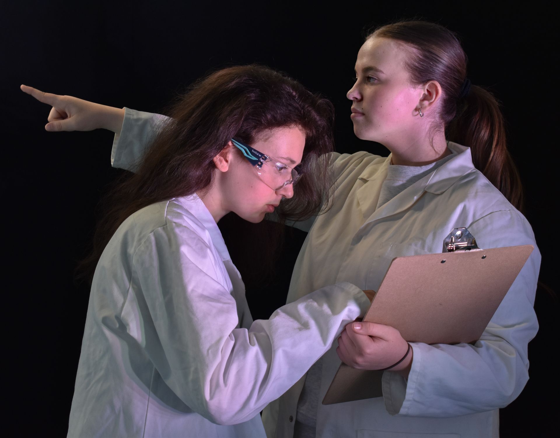 Two teenagers in lab coats reviewing a clipboard while one points offscreen against a black background